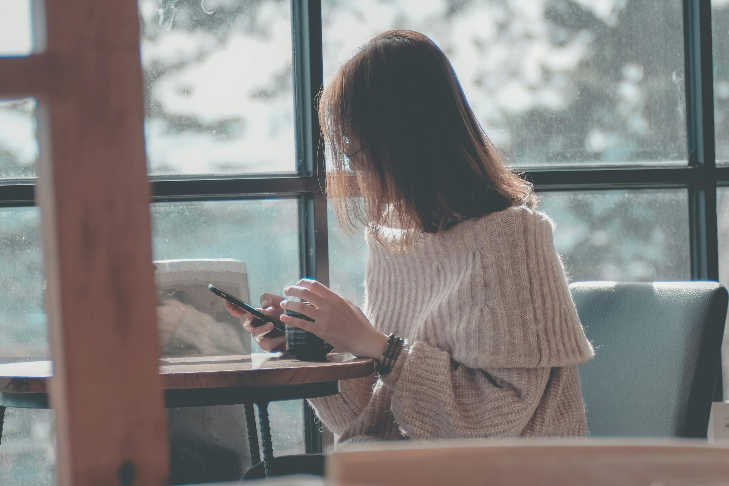 Woman using smartphone by a window with a view of trees, taking time off but communicating with her team. 