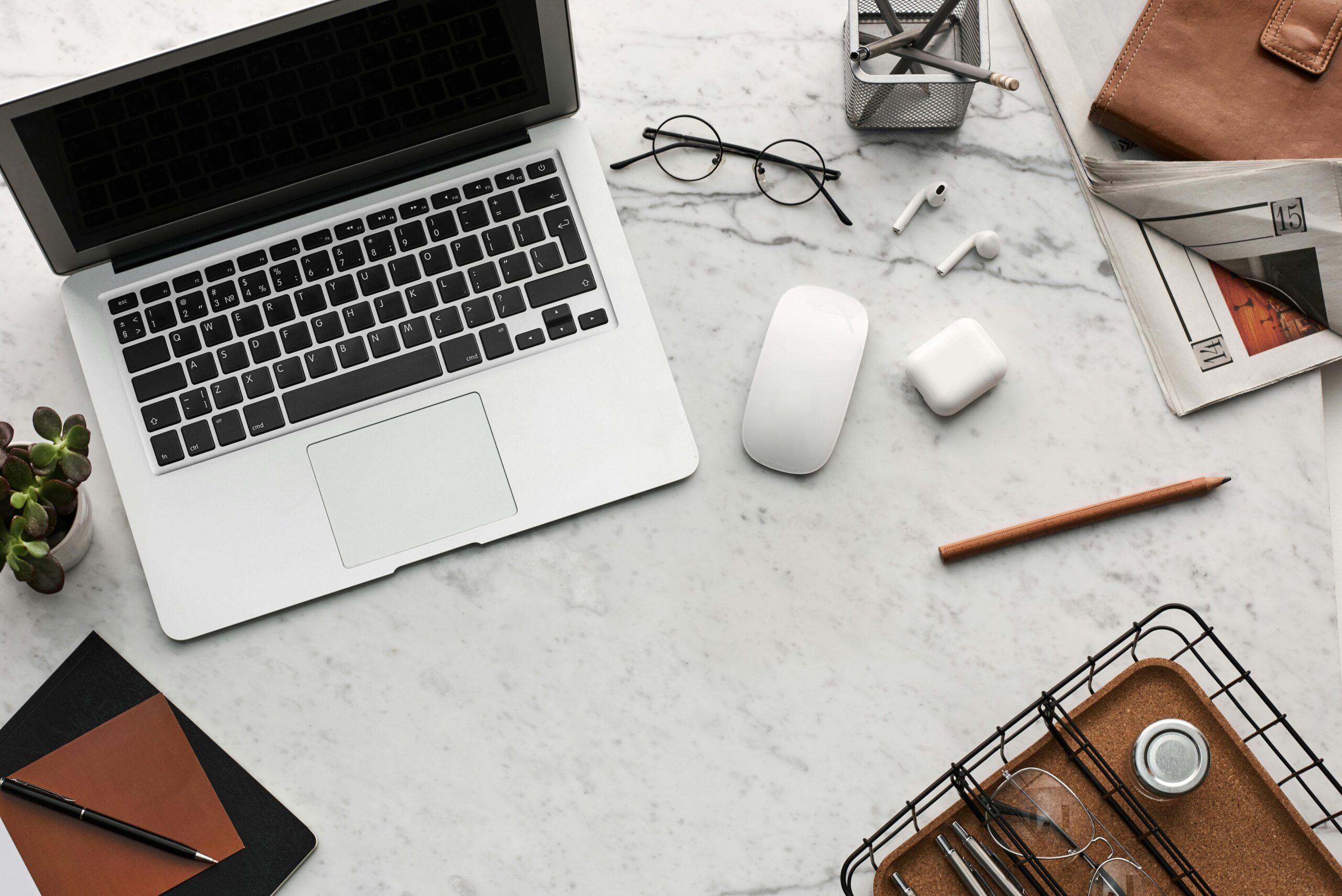 A neatly organized workspace with an open laptop, wireless mouse, eyeglasses, earbuds, notebook, and other office supplies on a marble surface.