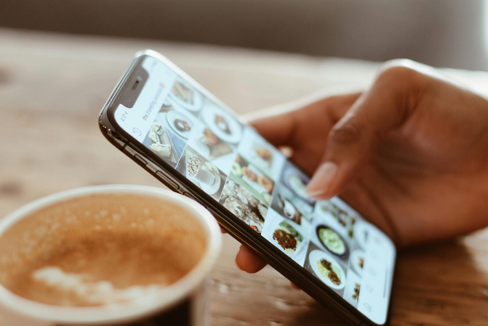 A person scrolling through social media a photo gallery on a smartphone beside a cup of coffee.