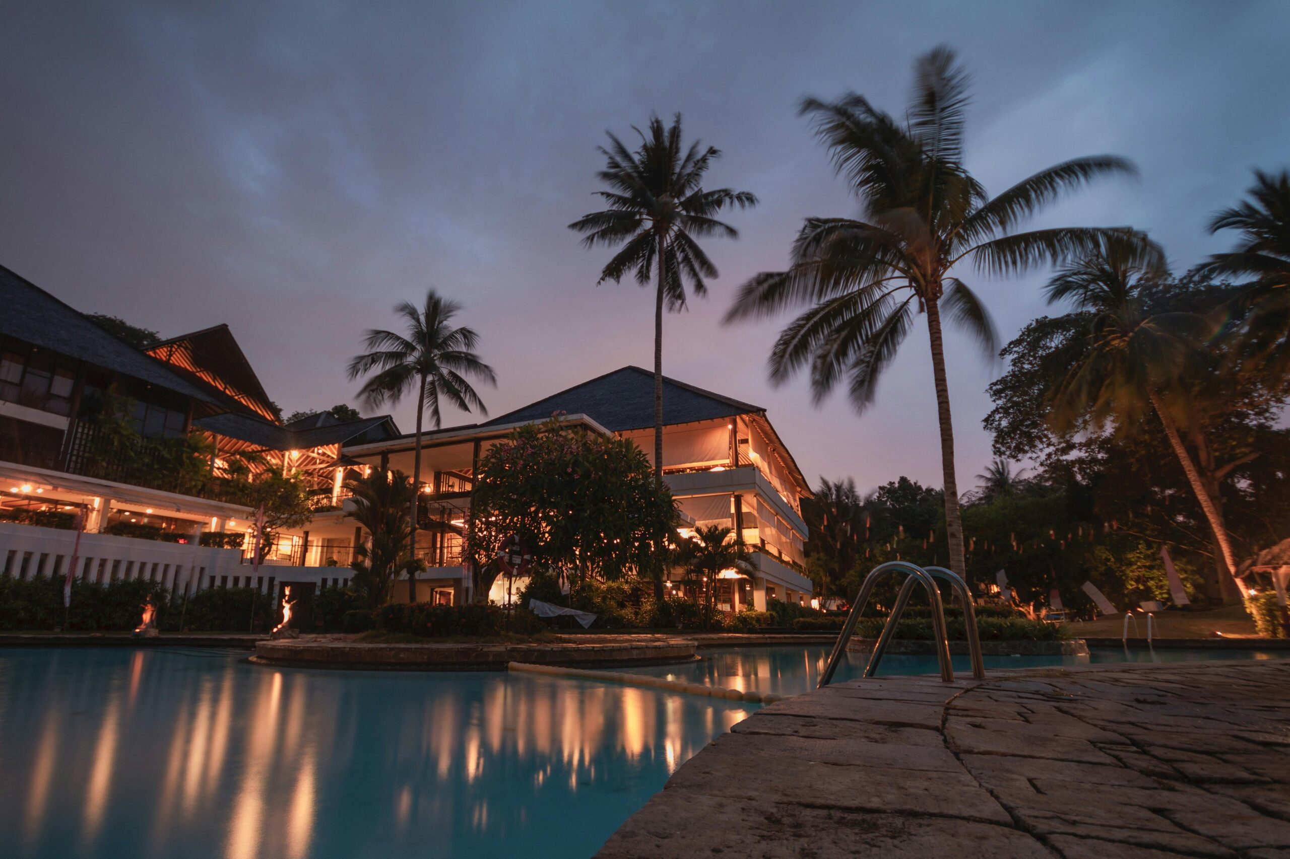 Tropical resort poolside at dusk with illuminated buildings and palm trees.