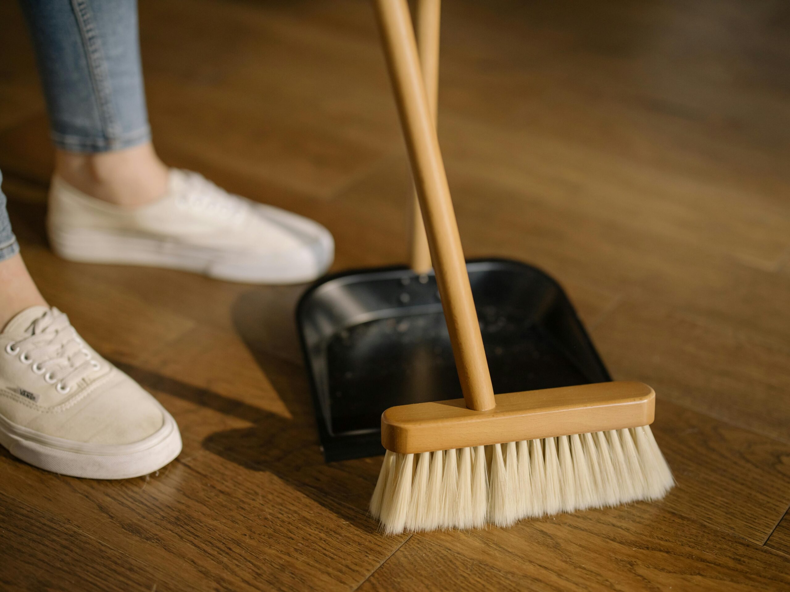 Person sweeping wooden floor with a broom into a dustpan, focusing on the lower half of their body and feet 