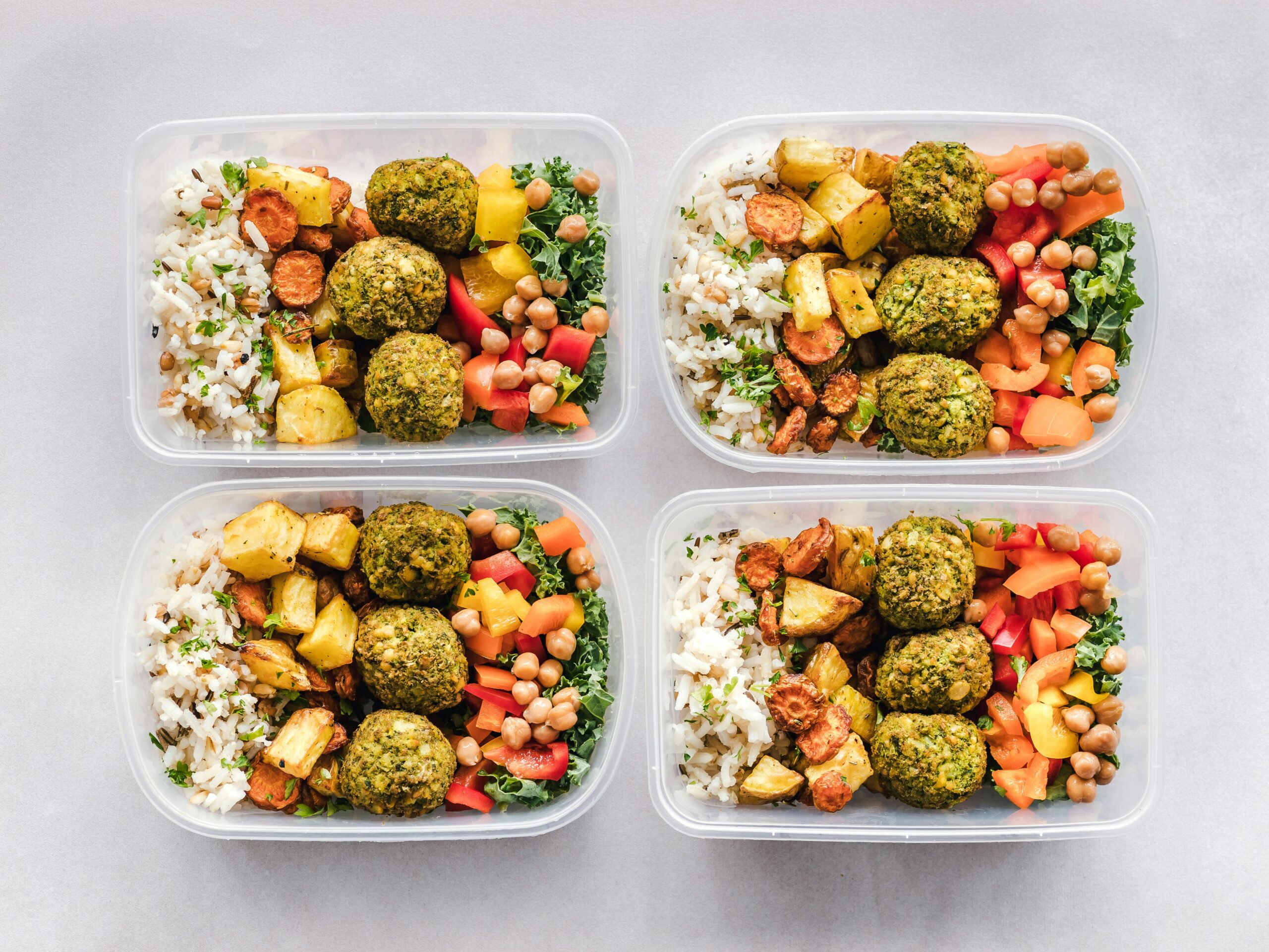 Four meal prep containers with assorted healthy foods including rice, chickpeas, vegetables, and falafel on a light gray background.