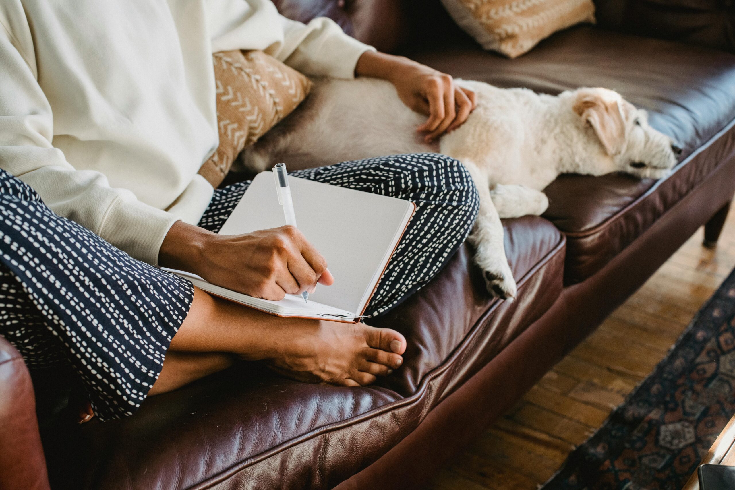 A person sitting on a leather couch writing in a notebook with a dog sleeping next to them working while being the own boss